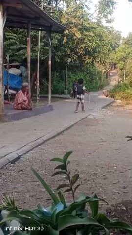 A child in a checkered shirt and shorts stands on a dirt path, holding a toy and facing an older person sitting on a stone bench under a covered structure. The older person is wearing a patterned shirt and is looking towards the child. Trees and foliage surround the area, and a slight incline is visible in the background. The sun appears to be casting long shadows, suggesting it is late afternoon or early morning. The scene appears to be outdoors in a rural or semi-rural setting.