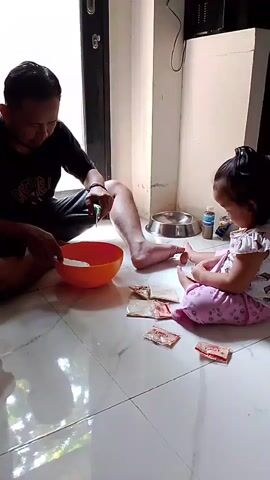 A man and a young girl are sitting on the floor. The man is stirring something in an orange bowl with a spoon. The girl is looking at some small packets on the floor in front of her. The scene appears to be indoors, possibly a kitchen or dining area, with tiled flooring.