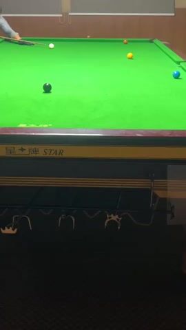 This video captures a game of snooker. A person, partially visible on the left side of the frame, is in the process of striking the cue ball with a snooker cue. The cue ball is positioned on the green snooker table, and the player's action is captured mid-stroke. Several colored snooker balls are scattered across the table, including a black ball, a white cue ball, a yellow ball, a red ball, and a blue ball. The snooker table has a characteristic green felt surface, and the surroundings appear to be an indoor game room, with the "STAR" brand name visible on the table's edge. The lighting is consistent with an indoor setting. The camera is positioned at a high angle, looking down at the snooker table.