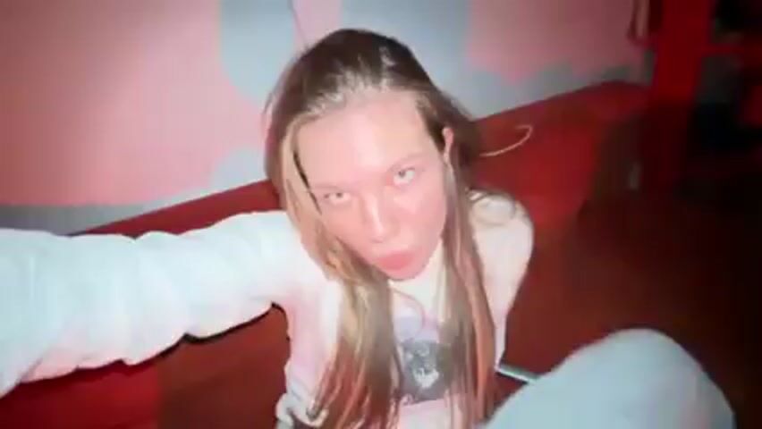 A young woman with long blonde hair takes a selfie-style video in what appears to be a bedroom. She is wearing a white long-sleeved shirt and white pants. The room has red and pink lighting. In the background, there is a large, round, colorful smiley face decoration on the wall, and a red couch. The woman moves the camera around to show the room and then makes silly faces at the camera. The music is playing in the background.