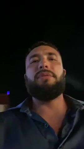 A man with a beard is smoking and looking around in the dark. He is wearing a blue shirt. The background is dark, with some blurred lights in the distance.