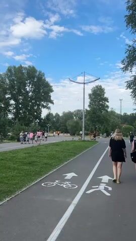 This media file captures a bright, sunny day in Abakan, Russia, likely late morning or early afternoon given the clear blue sky with scattered white clouds and the long shadows cast by the trees and lamppost. The scene is a park or recreational area, characterized by lush green grass, mature trees, and a paved pathway. The pathway is clearly marked with a white dividing line, separating a lane for bicycles (indicated by a bicycle symbol and arrow) from a lane for pedestrians (indicated by a walker symbol and arrow).

Several people are visible enjoying the park. On the left side of the pathway, a group of people, some walking and some possibly strolling, are moving along a grassy verge. Further in the background, other individuals can be seen, with a few people riding electric scooters. On the right side of the pathway, two young women are walking away from the camera, both dressed in casual summer attire. Their relaxed postures suggest they are enjoying a leisurely outing. The overall atmosphere appears peaceful and pleasant, typical of a summer day in a public park. There is a tall lamppost with a modern, curved design positioned near the center of the frame, and in the distance, there are hints of infrastructure like a tower, suggesting the park might be near an urban area. No visible text is present in the media.