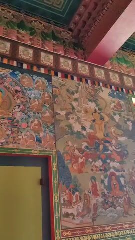 The video captures the interior of what appears to be a temple or monastery, characterized by ornate decorations and religious imagery. The camera pans across walls adorned with vibrant murals and intricate paintings. On the left side, a detailed mural depicts numerous figures, possibly deities or monks, arranged in a hierarchical manner around a central golden figure. The colors are rich, with prominent use of gold, red, blue, and green.

As the camera moves to the right, another large mural is shown, featuring a complex scene with multiple figures in various poses and settings, possibly illustrating a narrative or a religious story. These murals exhibit a traditional artistic style with fine details.

Further along the wall, a large collection of small golden statues, likely Buddhas or bodhisattvas, are neatly arranged in rows, creating a visually striking effect. The sheer number of these statues suggests a significant devotional or artistic undertaking.

The ceiling is also elaborately decorated with colorful patterns and symbols, including what looks like a Dharma wheel in one section. The architecture includes prominent architectural elements such as pillars and ornate borders. A large window with multiple panes lets in natural light, illuminating the polished floor. The overall environment conveys a sense of grandeur, reverence, and cultural richness, indicative of a sacred or historically significant space. There are no people visible in the video, and the focus is solely on the architectural and artistic elements of the interior.