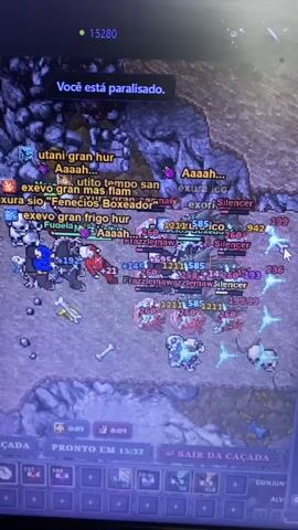 The video shows a screenshot of a video game, likely a role-playing game, displaying a chaotic battle scene. The environment is a rocky, purple-tinged terrain with scattered bones and some structures resembling ruins. Numerous player characters and enemies are engaged in combat, indicated by flashing damage numbers, spell effects like fire and ice, and various character names with associated numerical values. A prominent message at the top of the screen reads "Você está paralisado," which translates to "You are paralyzed," suggesting one or more players are incapacitated. The bottom of the screen shows game interface elements, including timers, experience points, and action buttons. The overall impression is one of intense, fast-paced gameplay.