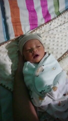 A newborn baby with dark skin is wrapped in a white and light gray blanket with a pattern of small dots. The baby is also wearing a light gray hat. The baby's eyes are partially open, and the mouth is slightly agape. The baby is held in someone's arm and lies on top of a blanket with a multi-colored striped pattern.