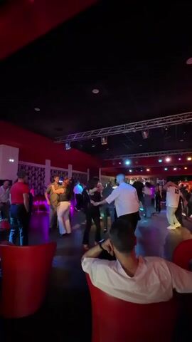 The video captures an indoor dance venue, likely a club or dance hall, with a lively atmosphere. Numerous people are engaged in couple dancing on a polished floor under dynamic, colorful lighting. Overhead, a truss system supports various spotlights and colored lights in shades of red, blue, green, and purple, illuminating the dance area. The ceiling is dark, and sections of the walls are red.

In the foreground, a person with short dark hair, wearing a white short-sleeved shirt, is seated comfortably in a red armchair, facing away from the camera, observing the dancers. Next to them, on a small table, a bottle and glasses are visible, along with a small sign or card that reads "carrone."

The dancers appear to be enjoying themselves, moving rhythmically, possibly to Latin music like salsa or bachata. The scene suggests an evening event, with the dim ambient lighting and vibrant stage lights contributing to a festive mood. The overall impression is one of social interaction and enthusiastic dancing in a dedicated dance space.