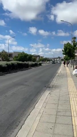 The video shows a street scene on a sunny day with a blue sky and white clouds. The camera is angled slightly upwards and to the right, capturing a road with traffic, a sidewalk, and buildings in the background. Several cars are driving on the road, and two people are walking on the sidewalk. There is a row of green bushes along the left side of the road, separating it from the buildings. Streetlights are visible along the sidewalk. The overall impression is of a moderately busy urban environment.