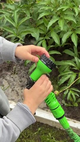 The video shows a close-up of a child's hands holding a green spray nozzle attached to a green hose. The child is rotating the nozzle head, which has multiple spray settings. Water is seen spraying from the nozzle. The background consists of green foliage and wet soil. The child is wearing a grey sweatshirt and yellow Crocs.