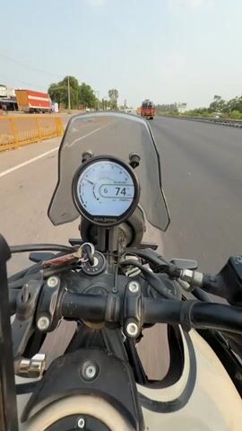 This media file captures a first-person perspective of a motorcycle ride on a highway near Panipat, India. The rider, wearing a red helmet and a black riding jacket, is moving at approximately 73-74 km/h, as indicated by the digital speedometer on the motorcycle's dashboard. The dashboard also shows the time as 05:14:45 and the odometer reading as 26889 km.

The scene is set on a multi-lane highway under a bright, hazy sky, suggesting it is either early morning or late afternoon. The weather appears to be warm and clear. The motorcycle is equipped with a small windshield.

Along the sides of the highway, there are occasional trees, poles, and buildings, including what appears to be a small roadside shop with yellow barriers in front. Traffic is light, with a few trucks and cars visible in the distance. The road surface is asphalt, and lane markings are clearly visible.

The rider has several small prayer flags attached to their motorcycle, adding a cultural element to the scene. The overall mood conveyed is one of steady, purposeful travel on a highway. The visible text includes "ROYAL ENFIELD" on the dashboard, the speed and time, and possibly some text on the prayer flags that is not clearly legible. The rider's face is not visible due to the helmet.