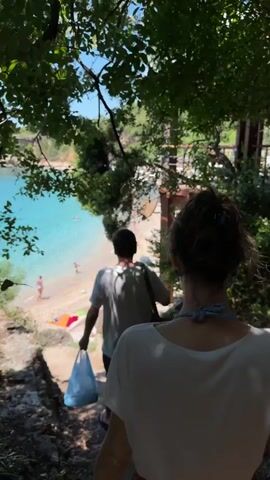 Two people are walking down a dirt path towards a beach. The person in front is a man wearing a white t-shirt and dark shorts, carrying a blue plastic bag. The person behind him is a woman, seen from the back, wearing a white shirt and a red skirt. The beach has a sandy shore and clear turquoise water. There are a few other people on the beach and in the water. The path is lined with trees and vegetation. The sky is blue and clear. The overall environment is a tropical or Mediterranean beach scene.