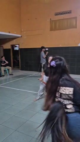 A group of people are inside a room with a green floor and yellow walls, possibly a gymnasium or community hall. In the foreground, a person with long dark hair is facing away from the camera, wearing a black t-shirt with a graphic design and blue jeans. They have a large bunch of long, light-brown hair or tassels attached to their back. To their left, another person is facing towards the camera, holding what appears to be a wooden prop with a shield-like attachment. They are wearing a black t-shirt and light-colored pants with a patterned design. In the background, several other individuals are visible, some holding similar hair or tassel props and dressed in casual clothing. One person in the background is holding a camera or phone, seemingly recording the scene. The overall atmosphere suggests a rehearsal or performance.