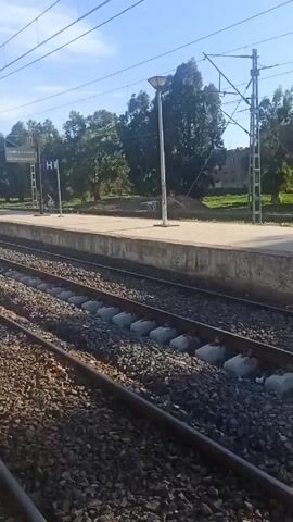 The camera pans along a railway platform, showing a sign reading "GARE MECHRAA BELKSIRI" in both Arabic and French. The platform is paved with patterned tiles and features a tactile paving strip for visually impaired people. The railway tracks run parallel to the platform, filled with gravel. Overhead power lines span above the tracks. Trees line the background, and a few buildings are visible in the distance under a partly cloudy sky. A fence runs along the edge of the platform. No people are clearly visible in the video.