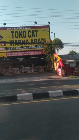 The video displays a street scene, potentially in Indonesia, viewed from a vehicle. A large yellow sign with red lettering dominates the foreground, advertising "TOKO CAT WARNA ABADI" (meaning "Eternal Color Paint Store"). The sign also includes details about opening hours and an address: "JL. SOLO-BOYOLALI KM. 22 WONOSARI, D.I. BOYOLALI". Electrical wires crisscross the sky above. To the right of the sign, a paved road with a distinct black and white curb is visible. Further down the street, trees and buildings are present, including what appears to be an office building with a sign that reads "KANTOR SEKRETARIAT IDI CABANG BOYOLALI" (meaning "IDI Branch Secretariat Office Boyolali"). The lighting suggests it is daytime, possibly late afternoon given the warm hue of the sunlight. The camera angle is tilted, giving a slightly distorted perspective. There are no discernible people actively engaged in actions within the frame, though some figures appear to be inside or near the store.