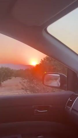 The video shows a view from inside a car. The camera is looking out the passenger side window and through the windshield. The time of day appears to be sunset, with the sky displaying hues of orange and pink. Outside the car, there are trees, bushes, and a dirt road. The road is visible in the side mirror as well. The interior of the car is dark and features part of the dashboard, a door handle, and air vents. The video captures a sense of movement as the car appears to be driving on the road through a rural, possibly hilly, landscape. The video is short, approximately five seconds long.