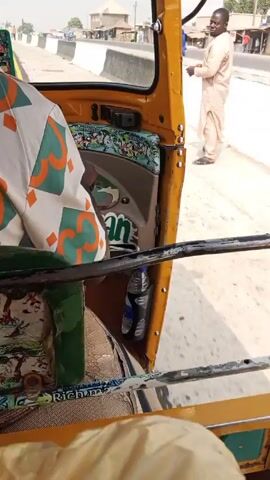 The video shows a view from inside a yellow tuk-tuk traveling on a paved road. The road has a yellow line marking the edge. The passenger is wearing a white patterned shirt. Outside, various vehicles are driving in the same direction. There are buildings and people visible alongside the road. The day appears bright and sunny.