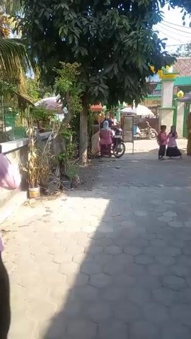A person wearing a black hijab and pink shirt walks away from the camera, her black skirt flowing. She is on a paved walkway with hexagonal stones. Behind her, two young children in pink shirts and dark skirts stand next to a woman in a pink headscarf sitting on a motorcycle. Further back, a green and yellow building is visible, possibly a mosque, with a palm tree and other foliage. The scene appears to be outdoors during the daytime, with sunlight casting shadows on the ground. The person in the black hijab appears to be moving towards the children and the motorcycle.