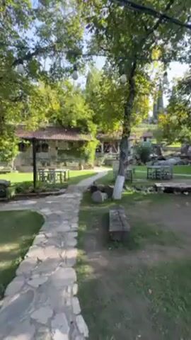 The video shows a view of an outdoor restaurant or gathering space. A stone pathway leads through a green grassy area dotted with trees, some of which have been painted white at the base. The space is decorated with various seating arrangements including tables and benches, as well as decorative items such as large clay pots. In the background, traditional style buildings with red-tiled roofs can be seen. People are milling around, suggesting a social gathering or a restaurant in operation. The scene appears to be set on a sunny day.