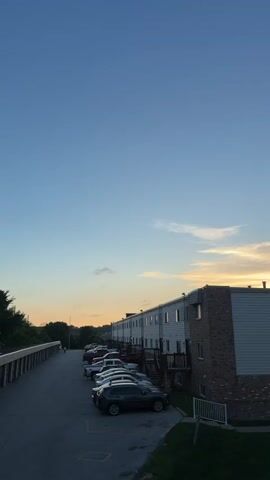 A low-angle, vertical shot captures a street with parked cars and apartment buildings under a clear blue sky. The sun is setting, casting a warm glow on the clouds and the tops of the buildings. A few wispy clouds are visible in the sky. The apartment buildings are two stories high and have a mix of brick and siding. There is a sidewalk running along the street and a grassy area in front of the buildings. The cars are parked in a lot next to the sidewalk. The video appears to be taken at dusk.
