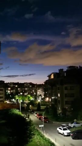 The media file shows a nighttime view of a residential area in Romashkovo, Russia. The scene is dominated by apartment buildings, illuminated by streetlights and the lights from within the apartments. Several cars are parked on the street and in a lot. The sky is a deep blue, with scattered clouds that are illuminated by an unseen light source, possibly from the setting sun or artificial lights on the horizon. The overall mood is calm and serene, typical of a quiet evening in a suburban area. There is no visible text in the media file.