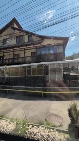 The video shows a series of shots taken from a moving vehicle, likely a car or bus. The scenery consists of buildings, streets, and vegetation. The first shot shows a multi-story building with a wooden facade and balconies. The second shot features a utility pole with numerous wires and a residential building in the background. The third shot displays a pink building and a white car parked on the street, with several people walking on the sidewalk. The fourth shot captures a wooden building with a sign for "Husqvarna" and a silver car parked in front of it. The final shot reveals a residential building surrounded by trees and greenery. The overall impression is that of a residential area or a small town.