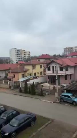 The video shows a residential neighborhood on a cloudy day. Several houses with red tile roofs are visible, along with a few cars parked on the street. Bare trees are scattered throughout the area.