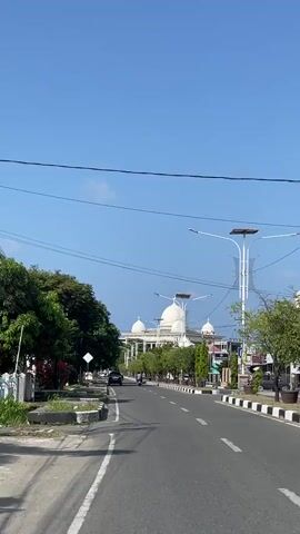 The video shows a view from a moving vehicle traveling down a paved road. The sky is clear and blue. On the left side of the road, there are trees and a sidewalk with a curb. On the right side of the road, there are more trees, sidewalks, and streetlights with solar panels. In the distance, a large white building with domes is visible, possibly a mosque or government building. Two motorcycles are visible on the road; one with two riders and another with one rider. A black car is also visible on the road. The overall scene depicts a sunny day in a town or city with developing infrastructure.
