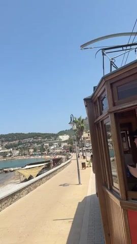 The video shows a waterfront promenade on a sunny day with a clear blue sky. To the right, the side of a wooden tram car is visible, with its windows reflecting the surroundings. The promenade is paved with light-colored square tiles and bordered by a stone wall. Beyond the wall, a beach with sand and pebbles stretches along the shoreline, with some beach umbrellas and lounge chairs. The sea is calm and blue. In the background, a coastal town with buildings is nestled on a hillside, and palm trees are scattered throughout the scene. A lamppost with a camera attached stands on the promenade, and a trash bin is also visible. Several people can be seen walking along the promenade and on the beach, and some boats are anchored in the bay. The camera appears to be mounted on the tram, providing a moving perspective of the seaside location.