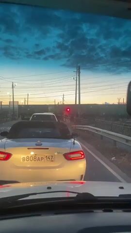 The video shows a view from inside a car driving towards a railroad crossing. The sky is a deep blue with scattered clouds, and the sun is low on the horizon, casting a warm glow. A white convertible car is in front, and a train is passing by on the tracks. The railroad crossing lights are red, indicating that traffic should stop. The car is stopped at the crossing, waiting for the train to pass.