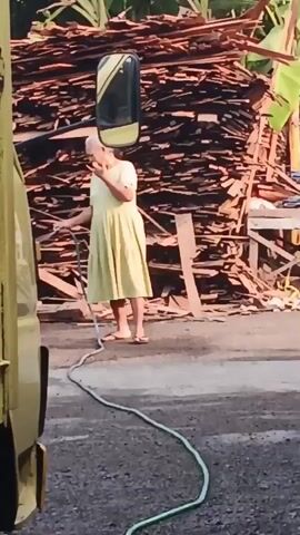 A woman in a yellow dress stands on a gravel surface next to a yellow truck. She holds a green hose in her left hand. A stack of wooden planks is behind her. A bicycle with a pink basket is visible in the background. The woman appears to be moving the hose.