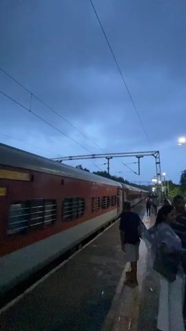 A train is stopped at a station platform in Sircaim, India. The train is red and grey with several windows. People are standing on the platform waiting to board or disembark. The sky is dark blue with some clouds, suggesting it is evening or early morning. There are overhead wires and lights visible on the platform. The overall atmosphere seems calm and ordinary, as one might expect at a train station.