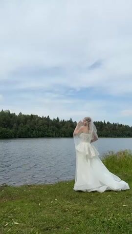 A woman in a white wedding dress and veil stands on a grassy shore, facing a body of water. Behind the water is a dense forest. The sky is cloudy. She is looking down and to her right, with one hand touching her veil. The video is very short, and the woman does not move significantly.