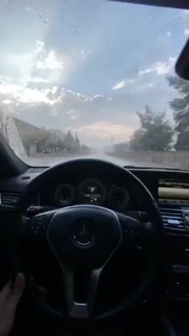 The video shows a view from inside a Mercedes Benz while driving on a wet road. It is raining, and the windshield is covered in raindrops. The car is moving slowly. The dashboard and steering wheel of the car are visible. The road is lined with trees and buildings. The sky is cloudy. The camera moves slightly during the video. The interior of the car is dark with light trim. There is music playing.