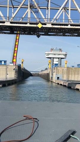 The video shows the perspective from a watercraft approaching a canal lock under a bridge. The lock is flanked by concrete walls and has a control tower with windows. A pedestrian walks across the canal further down the lock. A bridge spans overhead, supported by a steel structure. The water is slightly choppy. The sky is blue with few clouds. A yellow warning sign is visible on the bridge structure above the lock. A coiled rope lies on the deck of the vessel from which the video is being recorded.