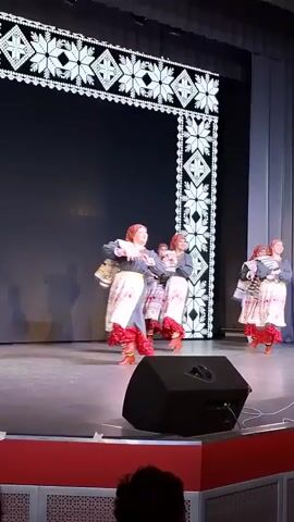 On a stage, six women in traditional Slavic folk costumes perform a dance. The costumes consist of grey blouses, red headscarves, and long white skirts with red trim. They also carry striped woven textiles which they twirl and toss during the dance. The stage is brightly lit, with a black backdrop adorned with white pixelated Slavic patterns. A large black speaker sits in the foreground. The floor is grey, with a red safety barrier at the edge of the stage visible in the lower portion of the frame. An audience member's head is visible at the very bottom of the shot.