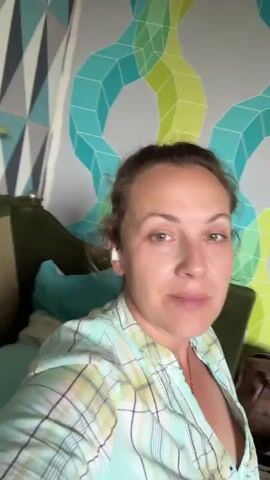 A woman with light brown hair pulled back is speaking directly into the camera. She is wearing a light green and white plaid shirt that is unbuttoned at the collar, revealing a necklace. She has on a small white earbud in her left ear. She is seated on a green couch, and behind her, the wall is covered with a geometric-patterned wallpaper in shades of gray, teal, and light green. A gold door handle is visible on the left side of the frame, on the wall. The lighting is neutral.