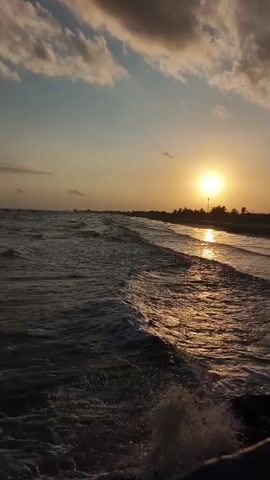 The video captures a sunset over the ocean. The sun is a bright yellow orb setting on the horizon, casting a golden glow on the water and clouds. Gentle waves roll towards a dark, sandy beach. Silhouettes of trees and possibly buildings line the distant shore. The sky is a gradient of orange, pink, and blue, with fluffy clouds scattered across it. The overall mood is serene and peaceful.