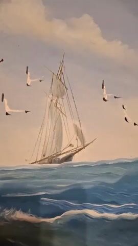 This is a close-up, low-angle video of an oil painting of a sailboat on the ocean. Seagulls are flying in the sky. The waves are blue and white. The sky is cloudy and a pale orange.