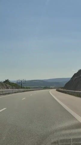 The media file shows a view from a vehicle traveling on a highway in a rural, mountainous region. The road is paved, with clear lane markings and guardrails on both sides. The scene is dominated by the road ahead, curving gently to the right, and the expansive, clear blue sky above. Rolling hills and mountains, covered in green vegetation, form the backdrop. The time of day appears to be late morning or early afternoon, given the bright sunlight and clear skies, suggesting fair weather.

On the left side of the highway, a large, empty billboard structure is visible. On the right side, a rocky, overgrown slope rises alongside the road. The overall impression is one of open space and travel through a natural landscape. There is no discernible text within the video itself, though the timestamp "00:00" and subsequent increments indicate the passage of time within the recording. The location context provided places this scene in Dzhurovo, Bulgaria.
