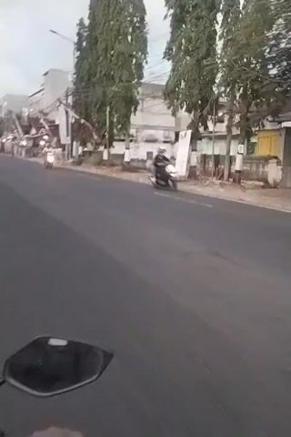 A first-person perspective video shot from a motorcycle shows a street scene. The road is asphalt with white lane markings. On the right side of the road, there are several buildings, some appearing to be shops or businesses. Some of these buildings have flags or banners displayed. Trees are visible along the side of the road. On the left side of the road, there are more buildings and some traffic, including other motorcycles and cars. The sky is overcast. The motorcycle appears to be moving down the street, with the handlebars and a mirror visible in the foreground.