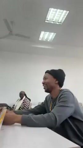 In a medium, low-angle shot, a man with dark hair and a beard sits in a classroom setting. He wears a black beanie, a grey sweatshirt with an indiscernible graphic, and a beaded necklace. He is speaking, gesturing with his hands, and appears to be engaged in a conversation. 

In the background, another person is visible, partially obscured, wearing a checkered top and head covering. Desks with books and papers are arranged in rows, typical of a classroom. The room has white walls and a perforated ceiling with recessed lighting panels. A ceiling fan is also visible. The overall lighting in the room is bright.