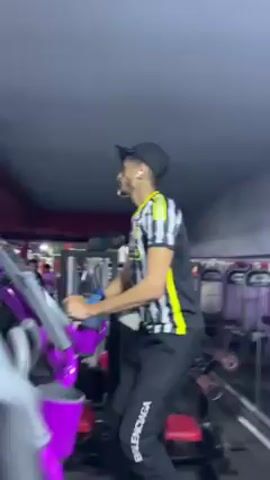 In a medium shot, a slender person in a black baseball cap, black and white striped shirt with yellow trim, and black pants with the word “Balenciaga” running down one leg, is using a stationary elliptical machine. The machine is largely purple. The person is facing away from the camera. In the background are other workout machines and people using them. The lighting is dim, with a purple hue.