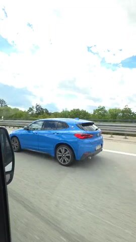 The video, shot from inside a moving vehicle, shows a blue BMW X2 driving ahead on a highway. The weather is sunny with a partly cloudy sky. The vehicle from which the video is taken seems to be a van or larger vehicle, as evidenced by the dashboard and the high viewpoint. A container of "Gazi" yogurt is visible on the dashboard. The highway is surrounded by trees. Other cars are visible in the distance. The camera briefly looks at the side mirror of the vehicle. The BMW maintains a steady speed.