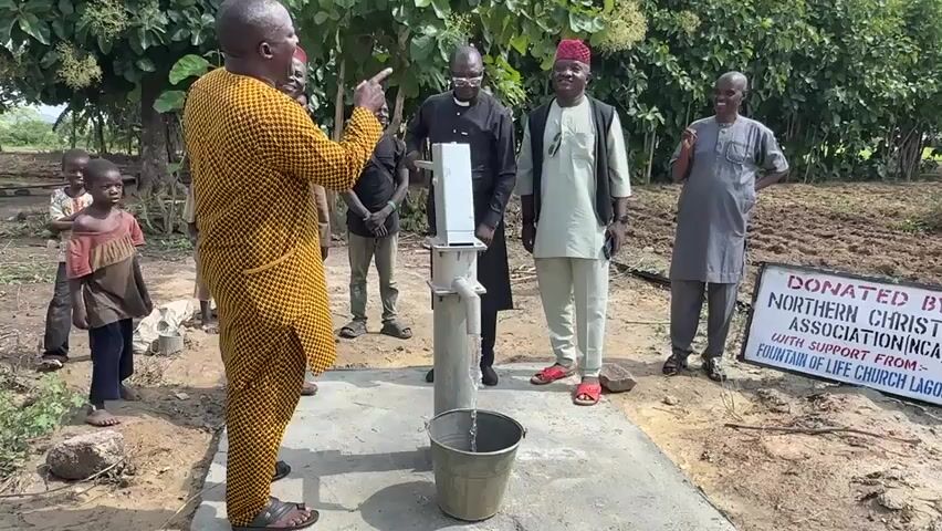 A group of people are gathered around a water well. A man in a yellow and brown patterned outfit is speaking and gesturing towards the well. A white water pump is attached to the well, and water is flowing into a silver bucket at the base. Several men and children are standing behind the well, observing. To the right of the well, a sign is visible, partially obscured by a man's leg, with text that appears to say "DONATED BY NORTHERN CHRISTIAN ASSOCIATION INC. WITH SUPPORT FROM FOUNTAIN OF LIFE CHURCH LAGOS". The environment appears to be outdoors, with trees and foliage in the background and a dirt ground surface. The overall impression is of a community event related to the installation or donation of a water source.