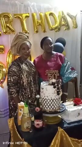 Two women stand behind a table decorated for a birthday celebration. The woman on the left wears a gold headwrap and a gold sequined dress. The woman on the right wears a maroon shirt with the words "Happy 50th" on it. Behind them, gold "BIRTHDAY" balloons are displayed. A cake decorated with black and gold accents sits on the table, along with two bottles of sparkling wine, a carton of juice, and other celebratory items. The backdrop is a white curtain. The setting appears to be an indoor party venue.