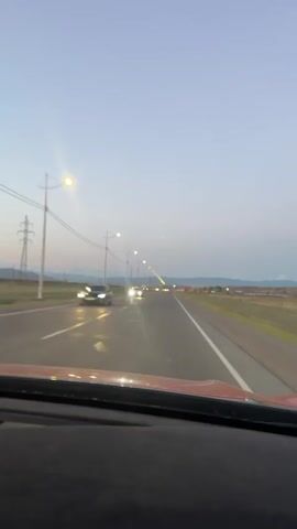 The video shows a view from inside a vehicle, looking out through the windshield at a road at dusk. The sky is a gradient of pale blue and purple. Streetlights line the right side of the road, illuminating the asphalt. There are power lines and poles also visible along the road. A few other vehicles are seen on the road ahead. The landscape to the sides of the road appears to be flat and covered in dry grass or vegetation. The vehicle is moving forward, as indicated by the slight blur of the scenery and the dynamic perspective.