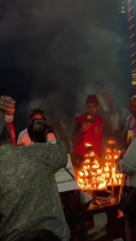 The video shows a group of people gathered around a decorative, tiered metal candelabra ablaze with numerous flames. Several people are holding up their phones, seemingly to capture photos or videos of the fire. One man in a red vest and a traditional hat is looking down at his phone. There is noticeable smoke rising from the candelabra. A man standing behind the candelabra is adjusting his face mask. The background is dark, suggesting it is nighttime, with a few illuminated decorations visible in the distance.