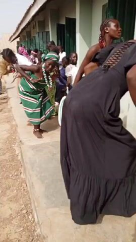 A group of young women dance outdoors in front of a light green building. The women are close to each other and wear a variety of casual clothes, including a long black dress and traditional West African patterned wraps and beaded necklaces. The camera is tilted, capturing the women’s movements.  The background includes green window shutters and a concrete surface. Loud music accompanies the dancing.