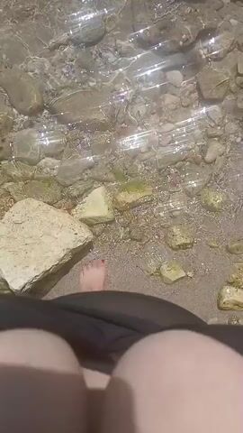 A first-person perspective video captures a person's feet standing in shallow, clear water over a sandy and rocky seabed. Sunlight glints off the water's surface, creating shimmering highlights. The rocks vary in size and color, with some appearing smooth and light-colored, while others are rougher and darker, some covered in green algae. The sandy bottom is visible beneath the water and in patches where the water recedes. The person's toes are visible, with red nail polish on the toenails of the left foot. A dark fabric, possibly part of clothing or a bag, is in the foreground, obscuring the rest of the person. The scene is peaceful and appears to be a natural outdoor setting, likely a beach or shallow coastal area. The time of day is suggested by the bright sunlight and the overall illumination of the scene, indicating daytime. No text is visible in the video.