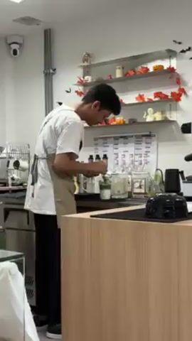 The video shows a young man in a cafe preparing a drink. The man is wearing a white t-shirt, black pants, and a beige apron. He is standing behind a counter in a well-lit cafe, meticulously adding liquid from a small metal container to a clear glass filled with ice and green liquid, presumably matcha.  The counter also holds various bottles and ingredients needed for drink preparation. Behind him, there is a shelf decorated with Halloween-themed items like pumpkins and ghosts.  A woman in a white shirt stands partially visible to the left of the barista. The environment appears clean and organized.