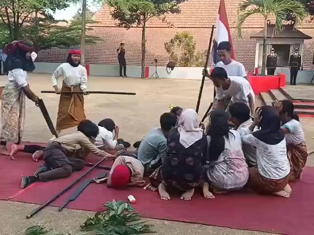A group of people are gathered outdoors, possibly for a performance or reenactment. In the foreground, a red mat is laid out on the ground. Several individuals are seated or kneeling on the mat, some appearing to be in distress. Two people standing on the left side of the mat are holding wooden staffs, and a third person on the right is also holding a staff and interacting with those on the mat. Scattered on the ground are more wooden staffs, and what appear to be imitation weapons. In the background, there are more people observing, some in what looks like school uniforms. A Dutch flag is visible on a flagpole. The environment appears to be an open area with some trees and buildings in the distance.